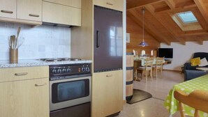 A kitchen with wooden cabinets, a stove, and a refrigerator. There is a dining area with a table and chairs, and a TV mounted on the wall. - Boden 19 by Interhome (Brienz)