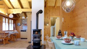 A cozy wooden cabin interior with a central wood stove, a dining area with a table set for four, and a kitchen area with wooden cabinets and a window. - Boden 19 by Interhome (Brienz)