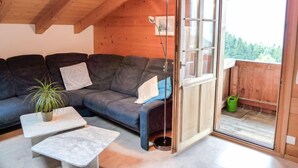 A cozy room with a dark gray sofa, a small white table, and a potted plant. A sliding glass door leads to an outdoor area with a wooden deck and a green bucket. - Boden 19 by Interhome (Brienz)