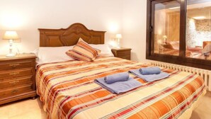 A hotel room with a wooden bed, striped bedding, a nightstand with a lamp, and a large window with a view of a stone wall. - La Tilia II by Interhome (Calonge)