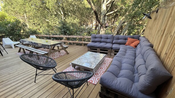 An outdoor deck with a wooden floor, a grey sofa, a small table, two chairs, and a wooden fence in the background.