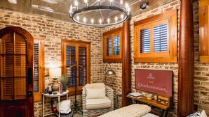 A room with a brick wall, wooden shutters, a chandelier, a chair, a table with a lamp, and a framed sign. - Luxurious Mansion Downtown Charleston (Charleston)
