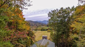 A cabin with a green roof, surrounded by autumn foliage and hills in the background.