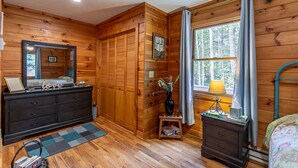 A wooden cabin bedroom with a dresser, a vanity, a bed, and a window with curtains.