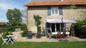 A two-story stone house with a tiled roof, a patio with outdoor furniture, and a garden.