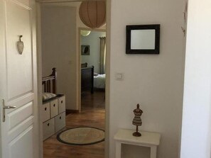 Un couloir avec un plancher en bois, une porte blanche, une petite table blanche ornée d’un objet décoratif et un miroir encadré accroché au mur.