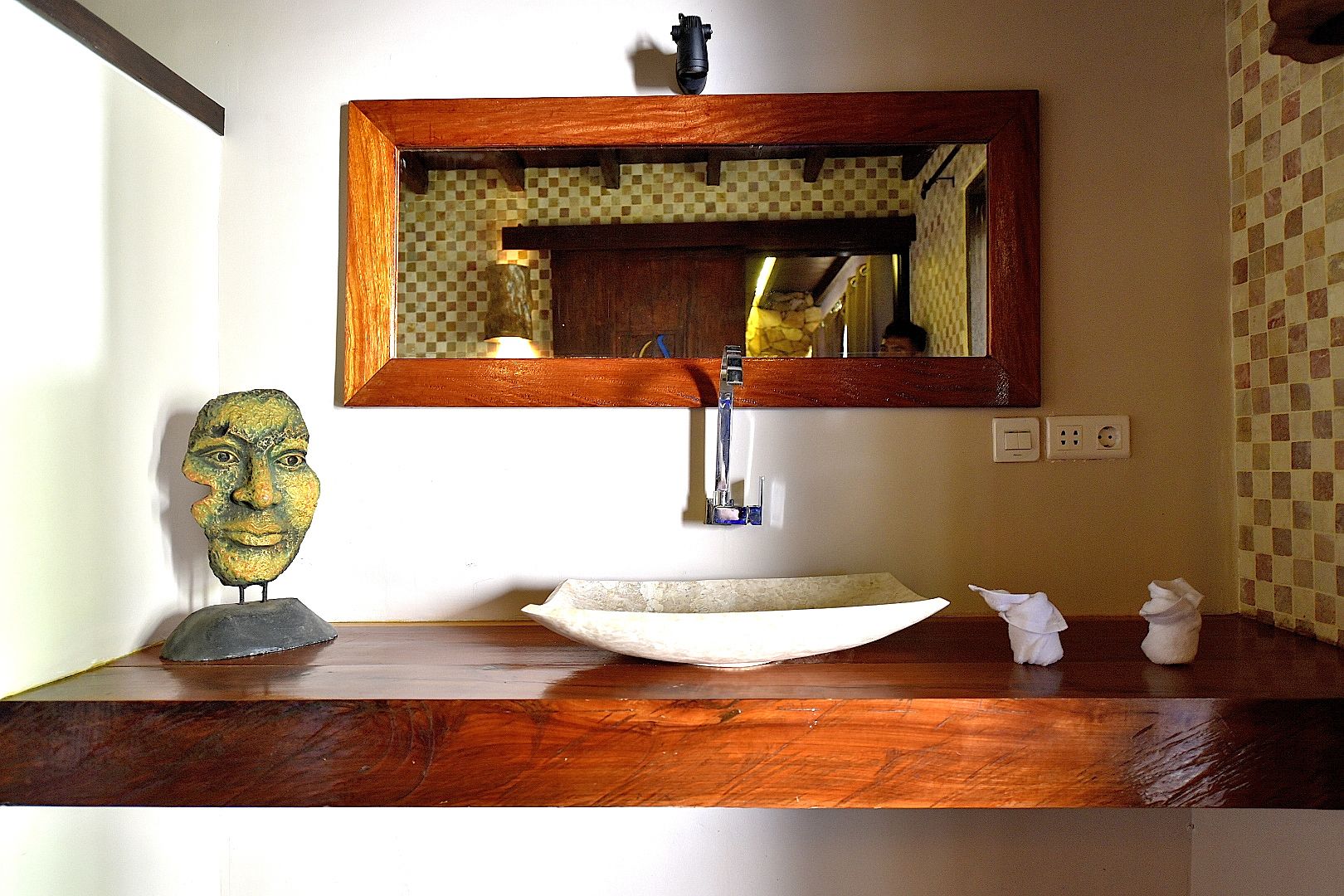 A bathroom with a wooden vanity, a sink with a vessel basin, a mirror, and a decorative head sculpture.