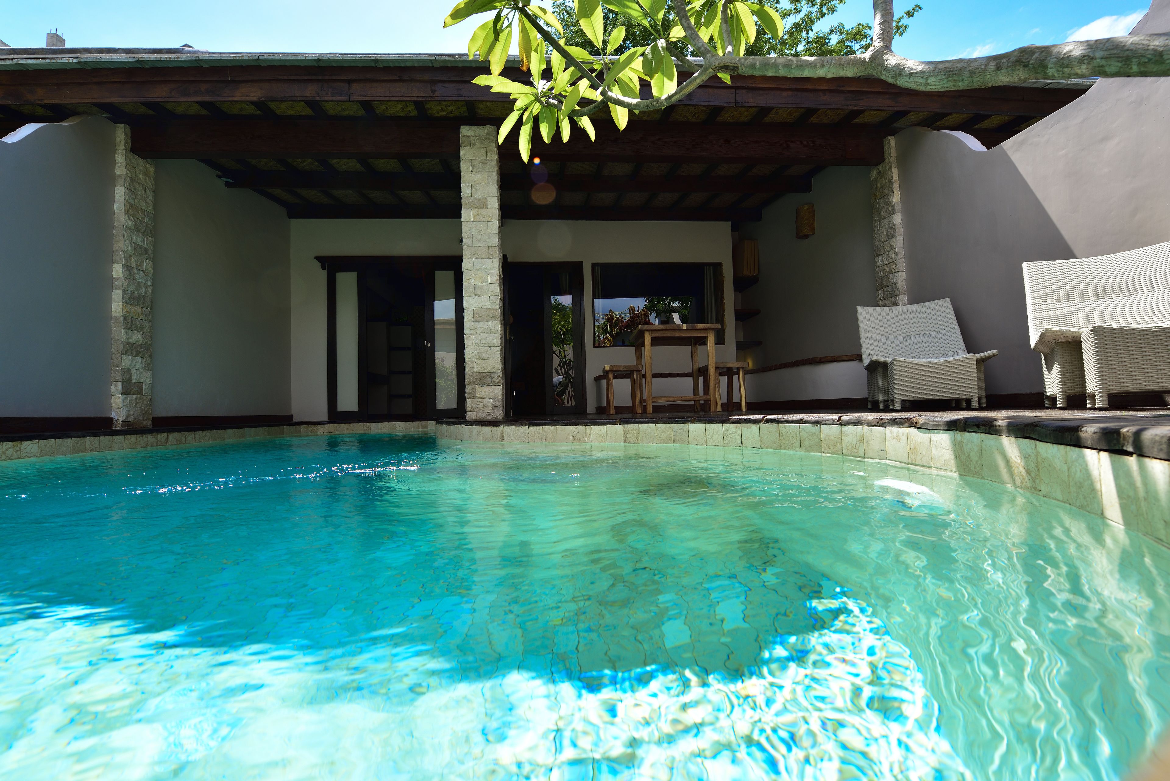 A swimming pool with clear blue water, a covered patio area with wicker chairs, and a stone pillar.