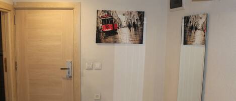 Quarto com porta de madeira, luminária fixada na parede e duas molduras com imagens de um eléctrico e uma cena de rua.