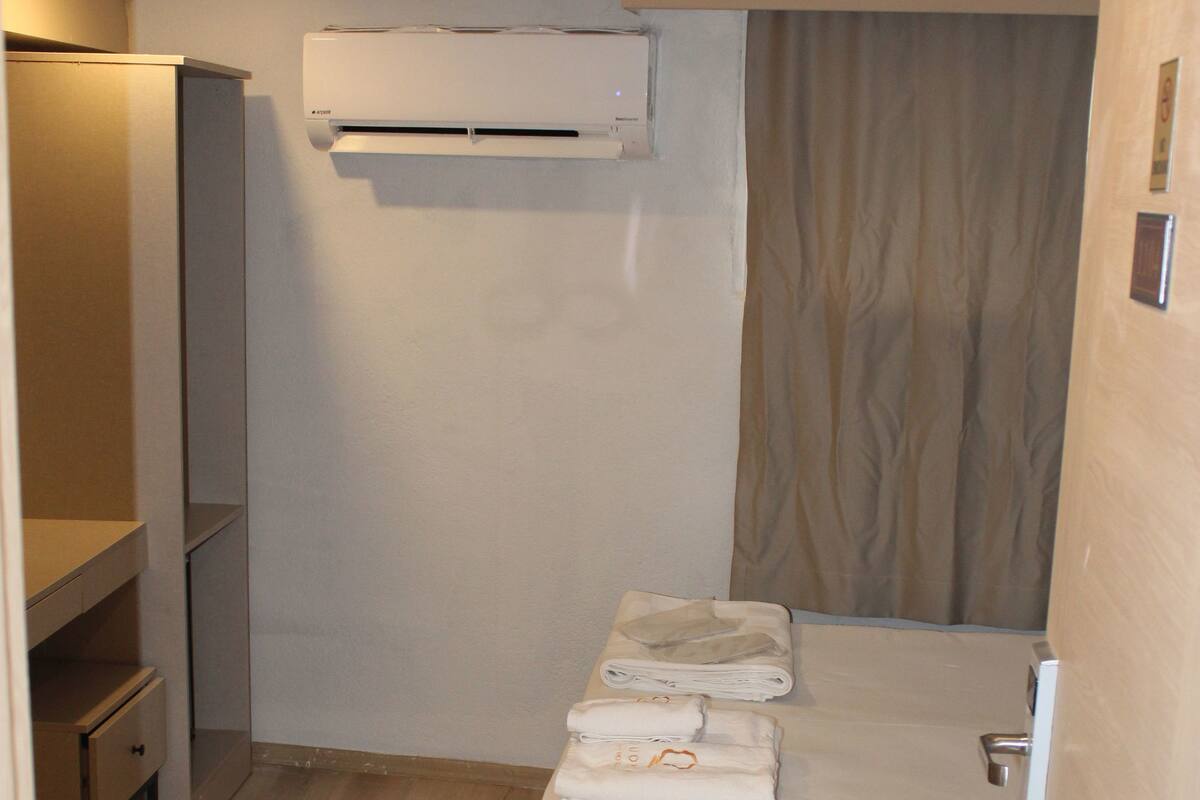 A small, compact room with a bed, a bedside table, a wardrobe, and an air conditioning unit mounted on the wall.