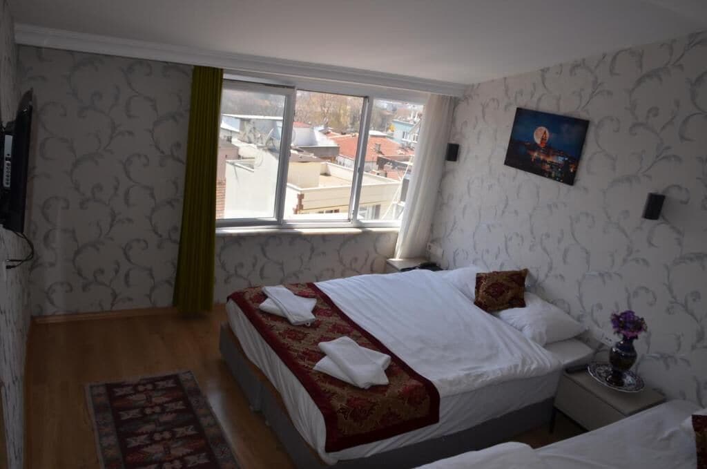 Une chambre d’hôtel avec un lit, une table de chevet, un vase de fleurs, une fenêtre donnant sur des immeubles et une affiche accrochée au mur.