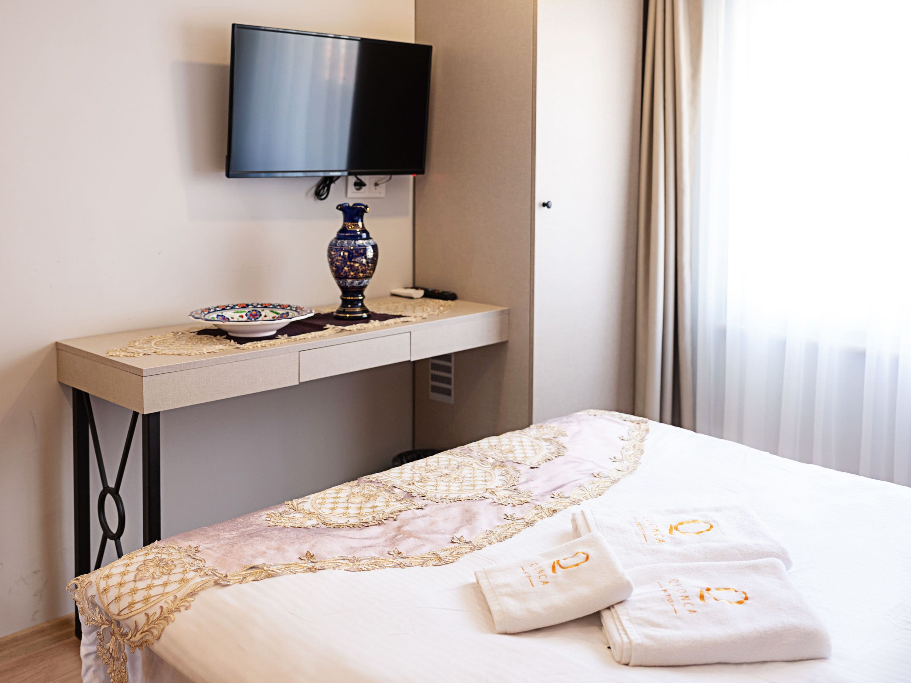 A hotel room with a bed, a TV mounted on the wall, a bedside table with a vase and a bowl, and a window with curtains.