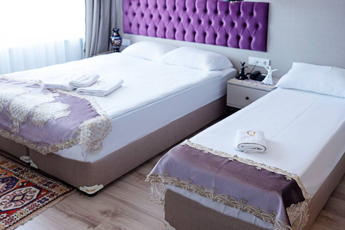 A hotel room with two beds, a purple headboard, white bedding, a nightstand, a lamp, and a decorative vase.