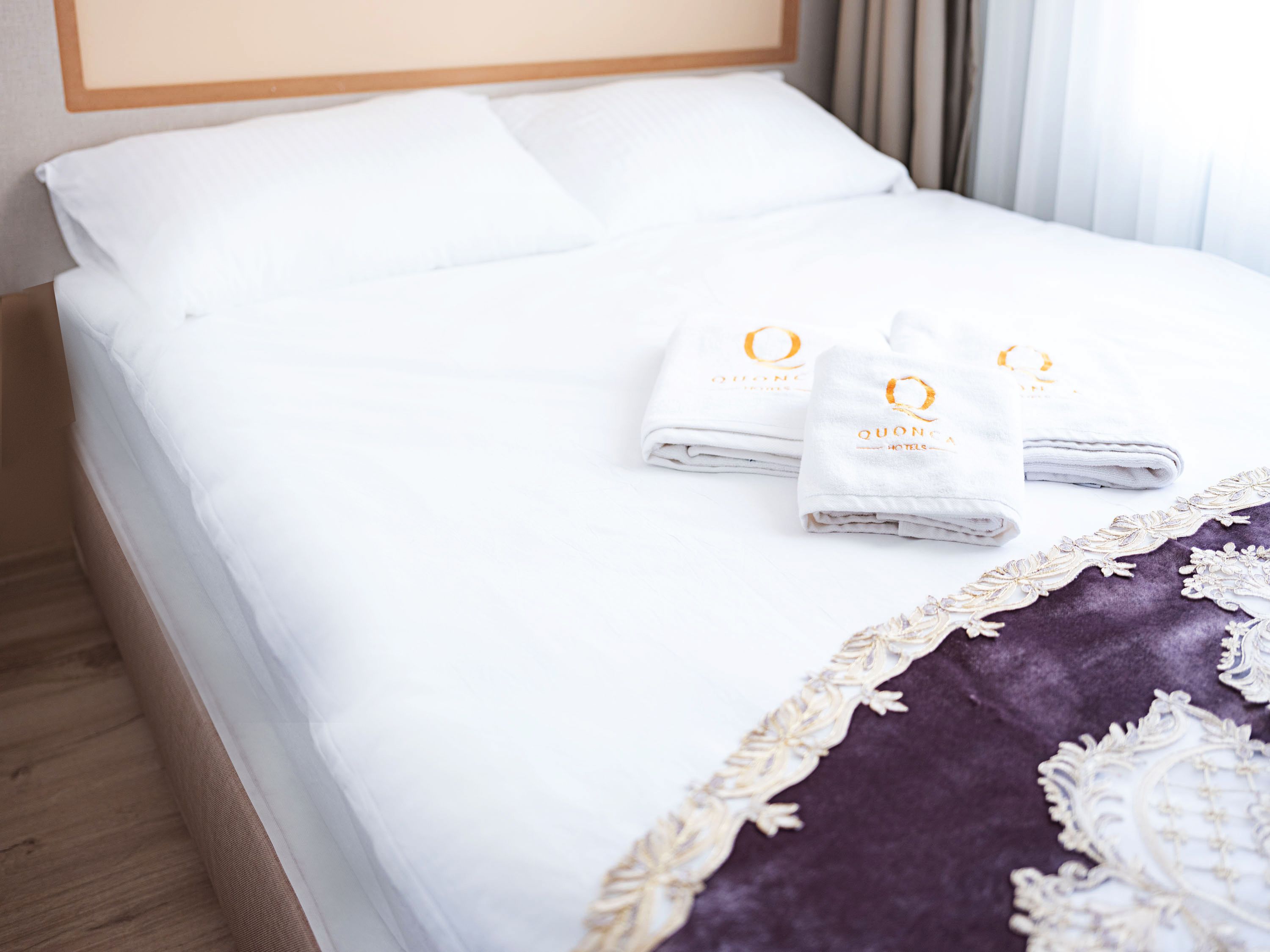 Un lit bien fait, avec du linge de lit blanc et une bordure décorative violette. On y trouve trois serviettes pliées, portant le logo d’un hôtel.