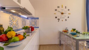 Una cocina con un reloj, un frutero y una zona de comedor con sillas y una mesa.