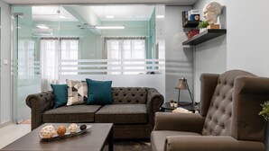 A modern living room with a grey sofa, a brown armchair, and a coffee table with decorative balls. There is a bookshelf with books and a bust on the wall. - Monastiraki Urban Apartments (Athens)