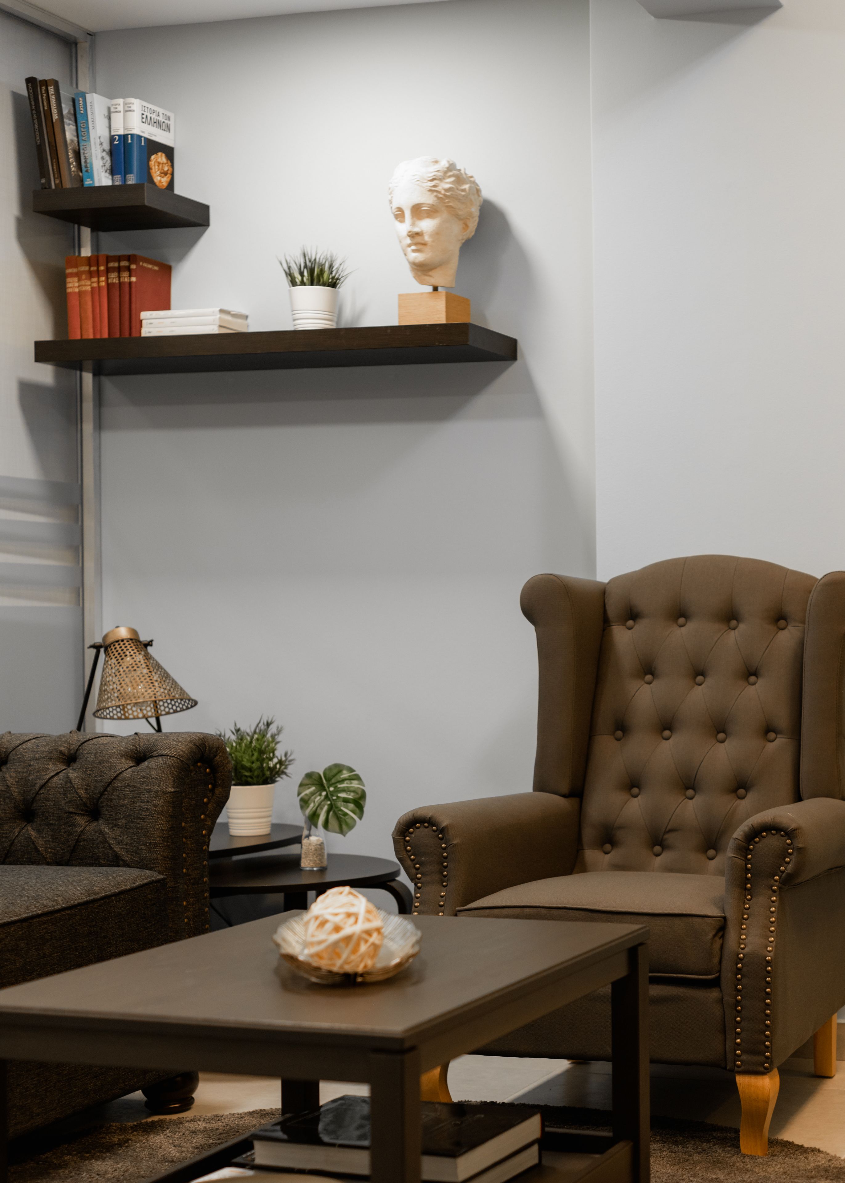 A living room with a brown armchair, a small table with a decorative item, a bookshelf with books and a bust, and a potted plant.