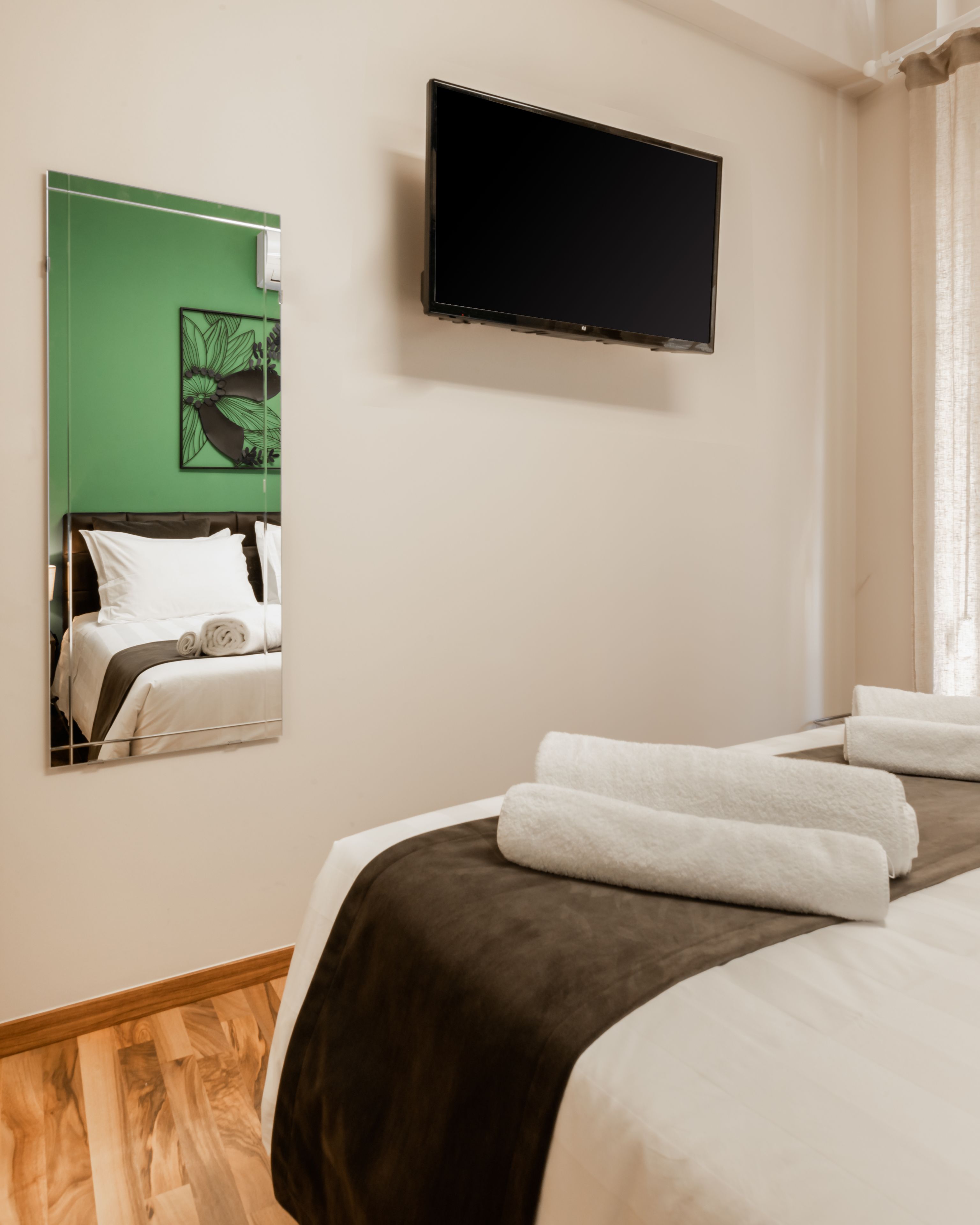 A hotel room with a bed, a flat-screen TV mounted on the wall, and a green-framed mirror reflecting the bed.