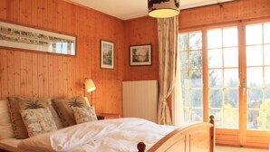 A wooden-paneled bedroom with a large bed, a nightstand, a lamp, and a window with curtains. - Armorial II / apt 2 by Interhome (Villars)