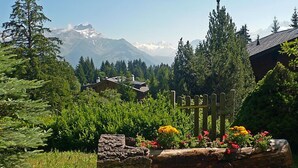 A mountain landscape with a wooden fence and colorful flowers in the foreground, and a house with a sloped roof in the background. - Armorial II / apt 2 by Interhome (Villars)