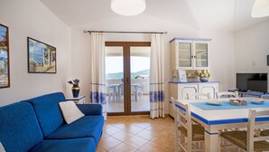 Un salon avec un canapé bleu, un coin repas avec une table et des chaises, une cuisine avec des placards et une vue sur l’extérieur, accessible par une porte vitrée.