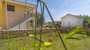Une aire de jeux avec des balançoires et des toboggans, un mur en pierre et un bâtiment doté d’un balcon.