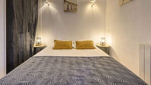 A bedroom with a patterned bedspread, two bedside lamps, a wall-mounted picture, and a curtain. - Océania II by Interhome (Carnac)