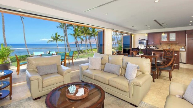 Uma sala de estar moderna com uma grande janela com vista para a praia, uma área de jantar com mobiliário de madeira e uma cozinha com armários escuros.