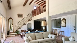 Un vaste espace de vie doté d’un plafond haut, de poutres apparentes, d’un escalier et d’un coin repas avec une table et des chaises.