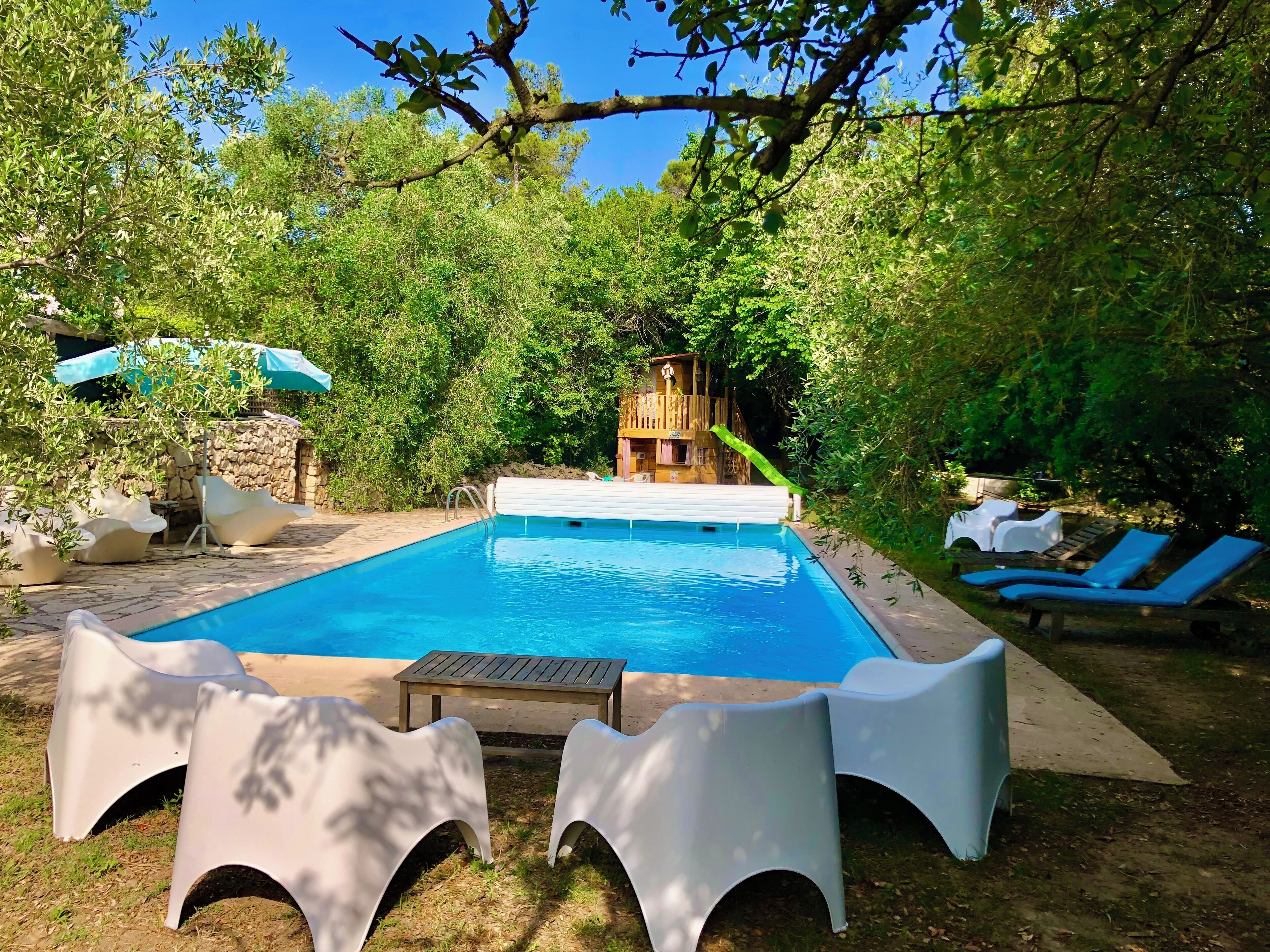 Une piscine dotée d’une terrasse en bois, entourée de verdure et bordée d’un mur en pierre.