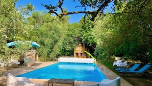 Une piscine dotée d’une terrasse en bois, entourée de verdure et bordée d’un mur en pierre.