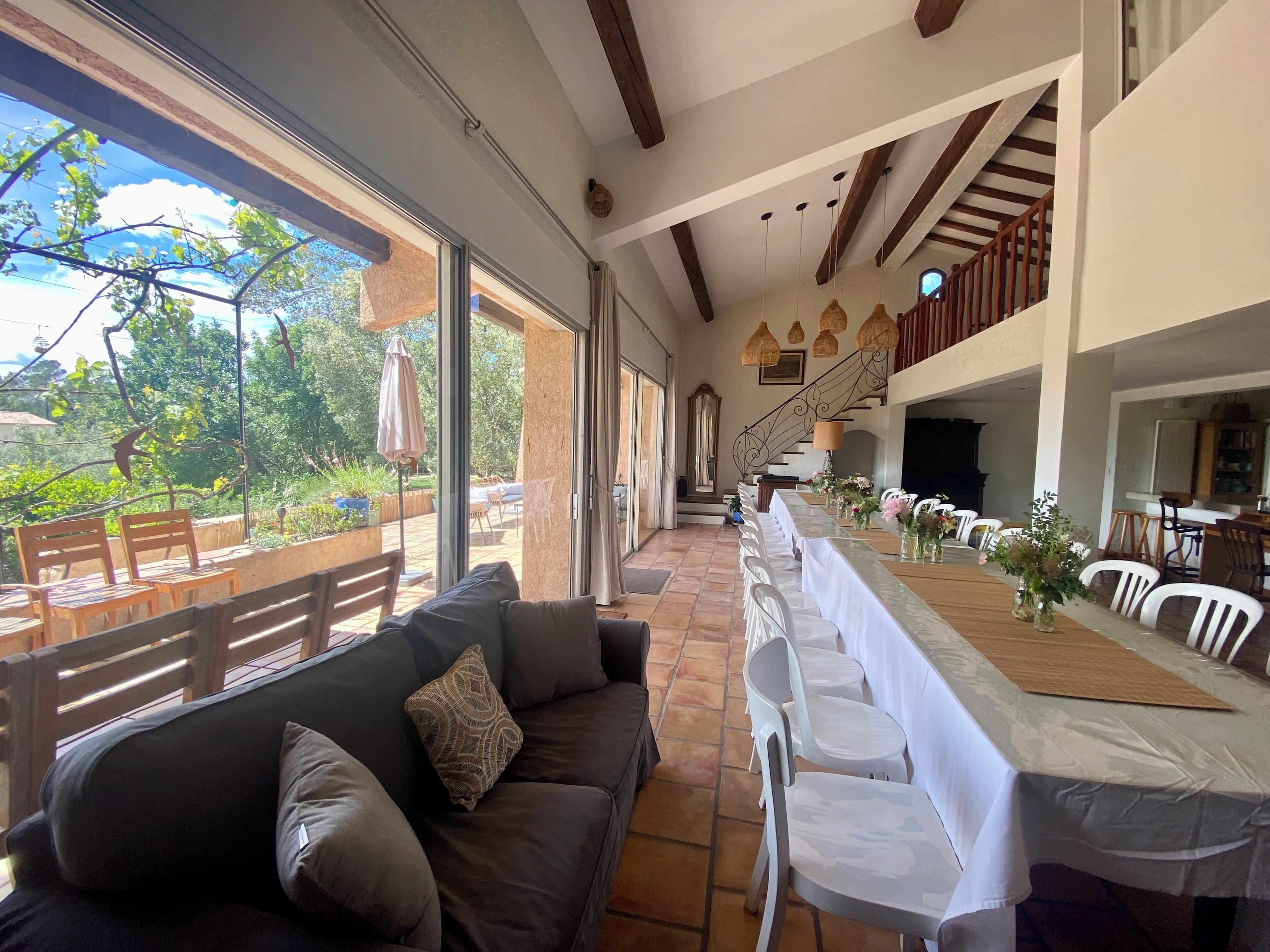 Un vaste espace intérieur comprenant une longue table à manger dressée pour un repas, un canapé avec des coussins et un escalier menant à un niveau supérieur.