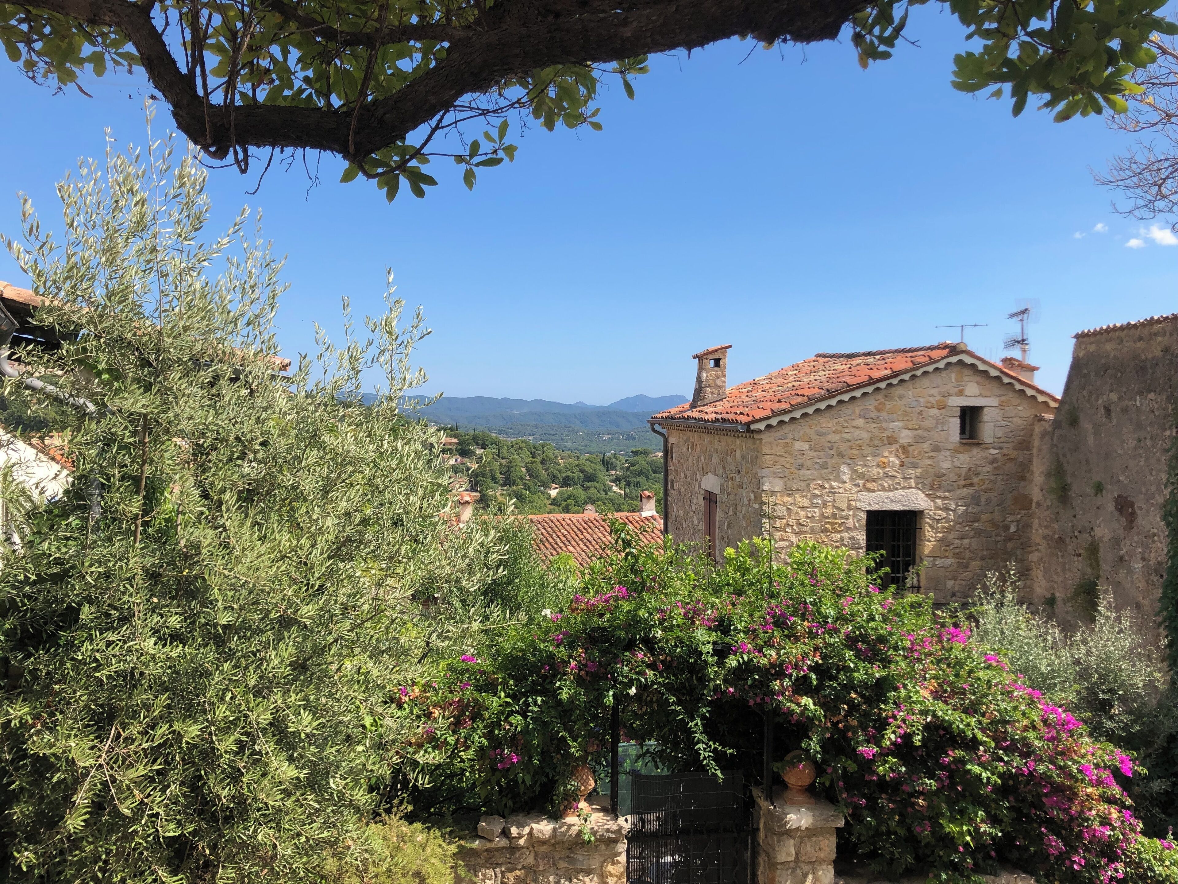 Un bâtiment en pierre, surmonté d’un toit en tuiles, entouré de verdure et offrant une vue sur des montagnes lointaines.
