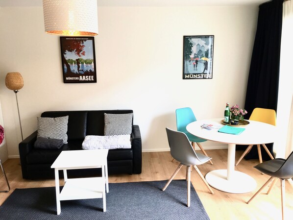 Ein modernes Wohnzimmer mit einem schwarzen Sofa, einem weißen Couchtisch und einem runden Esstisch mit Stühlen. An den Wänden befinden sich Poster und eine Lampe hängt über dem Sofa.