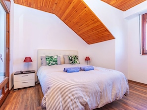 Un dormitorio con techo inclinado, una cama con ropa de cama a rayas, dos mesitas de noche con lámparas y una ventana.