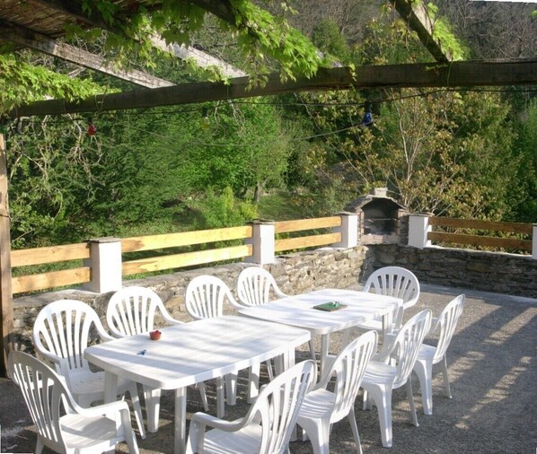 An outdoor patio with white plastic tables and chairs, surrounded by a stone wall and wooden fence, with a stone fireplace in the background.