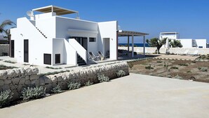 Un bâtiment moderne aux murs blancs, doté d’un mur de soutènement en pierre, d’un espace extérieur aménagé et couvert, et offrant une vue sur la mer.