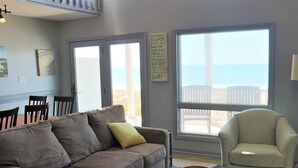 A living room with a brown sofa, a green armchair, a dining table with chairs, and a large window with a view of the beach.
