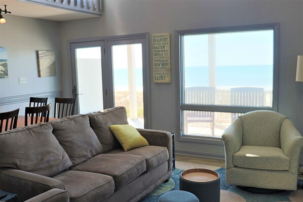 A living room with a brown sofa, a green armchair, a dining table with chairs, and a large window with a view of the beach.