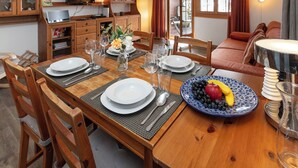Ein Essbereich mit einem Holztisch, gedeckt für vier Personen, umgeben von Stühlen. Eine Holzkommode mit einem Fernseher links und ein Sofa mit einer Lampe rechts.