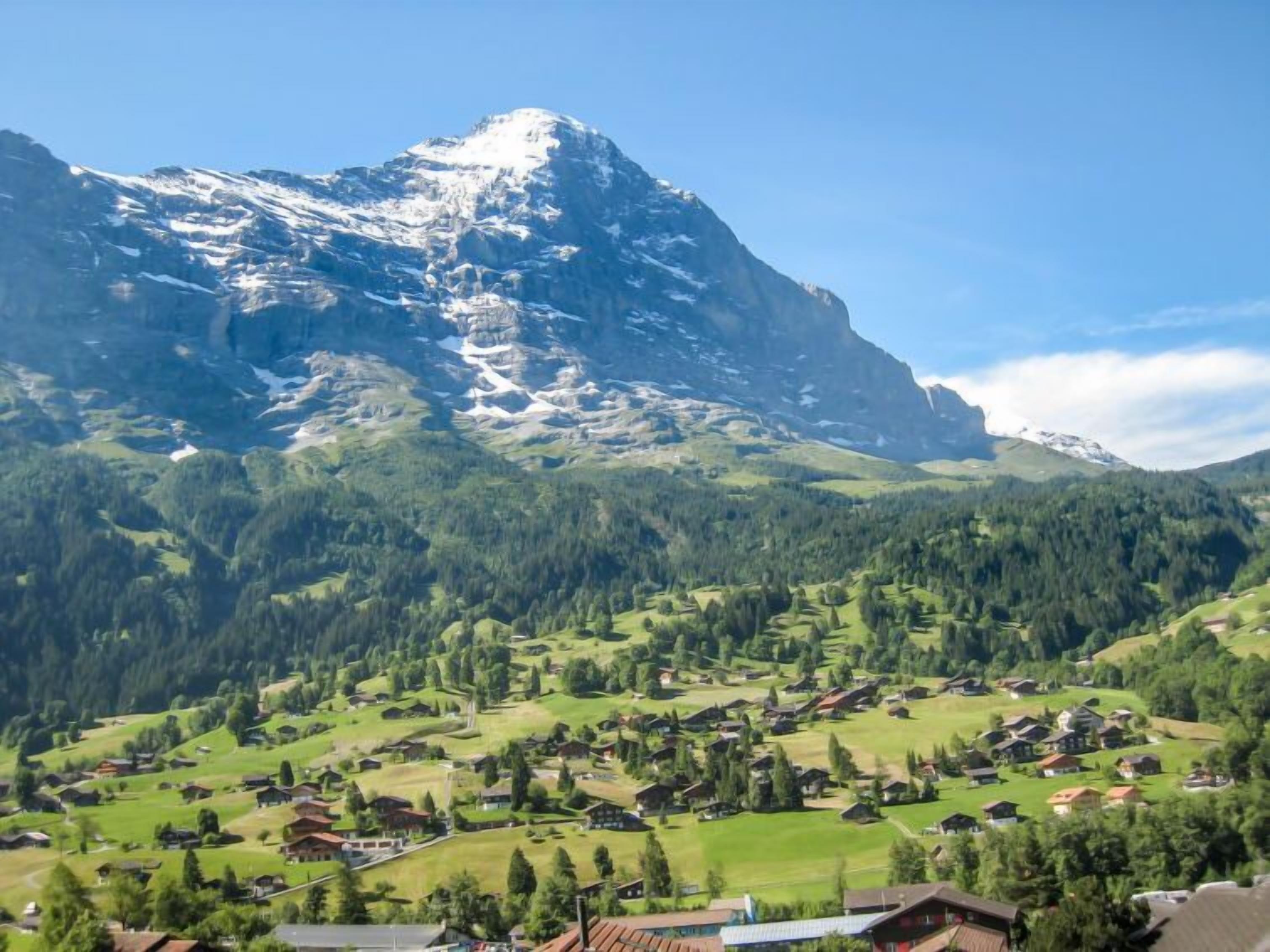 Un village de montagne aux maisons traditionnelles, sous un ciel dégagé, avec un sommet enneigé imposant qui se détache à l’arrière-plan.