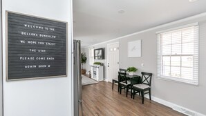 Um bistrô moderno com piso de madeira, área de refeições com mesa e cadeiras, e uma placa de boas-vindas na parede.