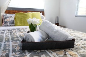 A neatly made bed with a floral-patterned bedspread, a black tray with folded towels, and a vase with white flowers.