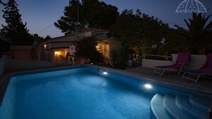 En swimmingpool med en kant av kakel, omgiven av solstolar och en liten byggnad med lampor.