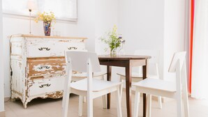 Un comedor con una mesa de madera blanca desgastada y sillas a juego, un jarrón con flores y una lámpara sobre un aparador blanco.