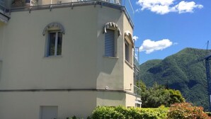 Un edificio en esquina con balcón, ventanas con contraventanas y montañas al fondo.