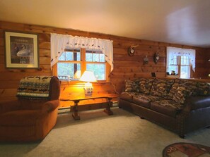 A cozy cabin interior with a wooden wall, a sofa, a chair, and a lamp near a window with curtains.