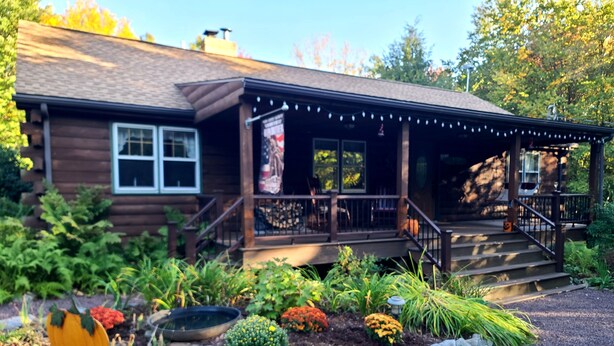 A wooden cabin with a porch, steps, and a garden with various plants and a pumpkin.