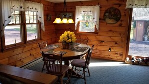 A wooden cabin interior with a dining table, chairs, a chandelier, and a window with curtains.