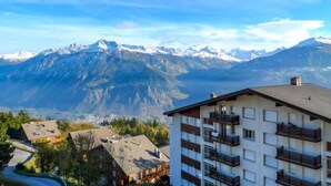 A multi-story building with balconies, set against a backdrop of snow-capped mountains and a clear blue sky.
